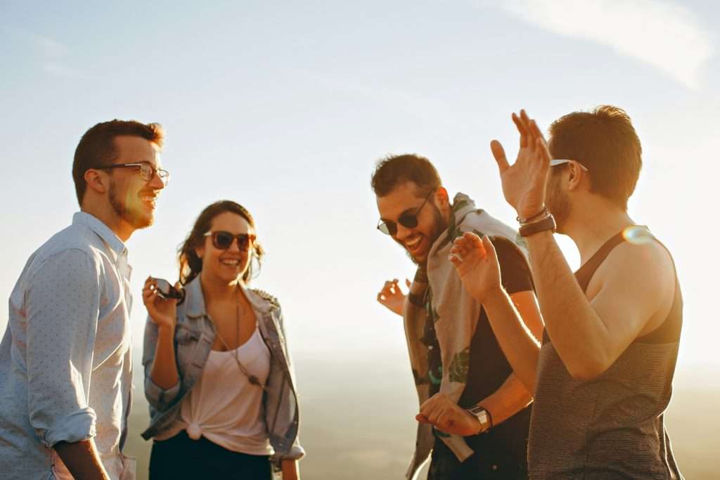 Four people are standing outside, smiling and laughing under a bright sky. One person is holding an object, while others have raised arms in animated conversation, clearly having fun. Their joy and camaraderie reflect the easygoing lifestyle of early retirement.