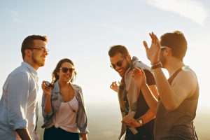Four people are standing outside, smiling and laughing under a bright sky. One person is holding an object, while others have raised arms in animated conversation, clearly having fun. Their joy and camaraderie reflect the easygoing lifestyle of early retirement.