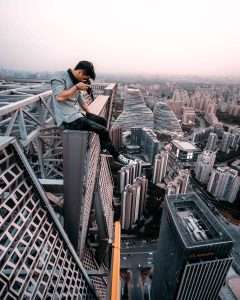 A person sitting on a high steel structure, capturing a photo with a cityscape in the background during dusk, perhaps dreaming of an early retirement amidst this extreme beauty.