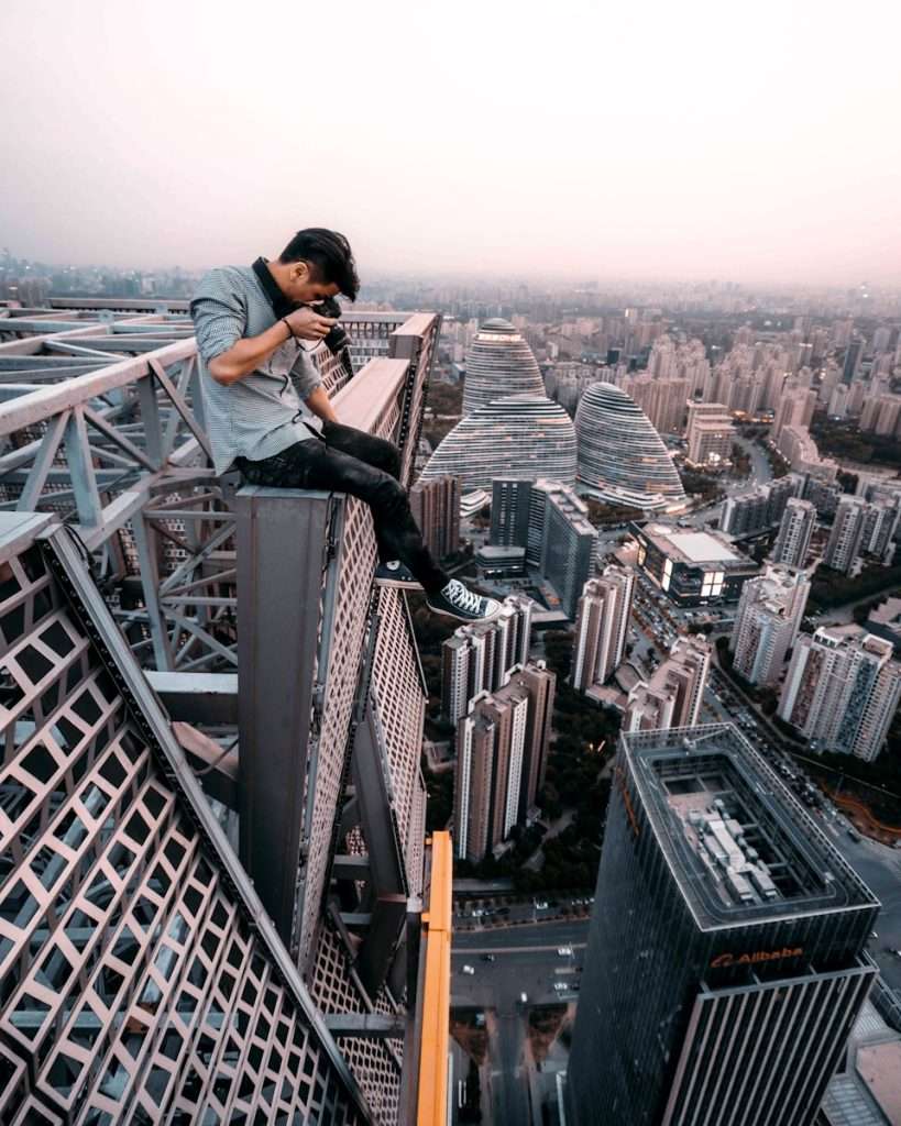 A person sitting on a high steel structure, capturing a photo with a cityscape in the background during dusk, perhaps dreaming of an early retirement amidst this extreme beauty.