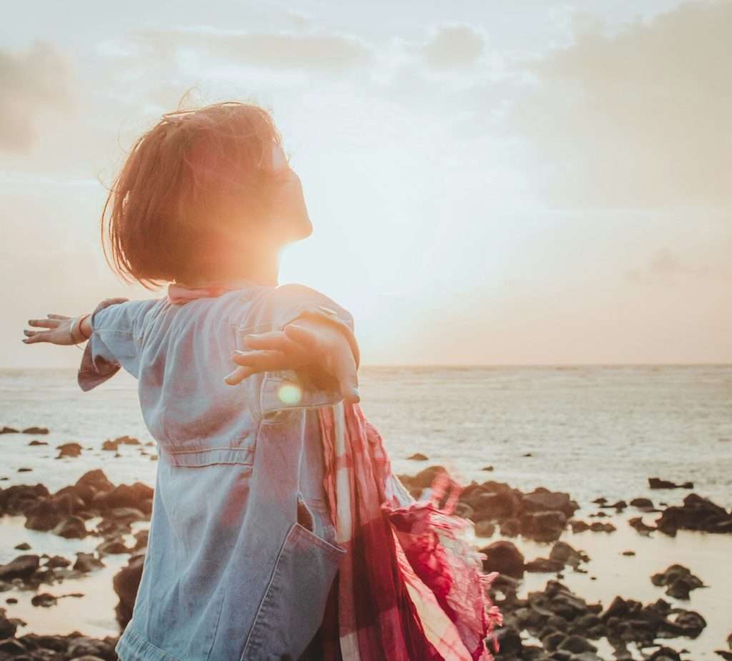 Person with short hair and open arms faces the sun at a rocky beach during sunset, with a scarf draped over one arm, unlocking joy in every moment.