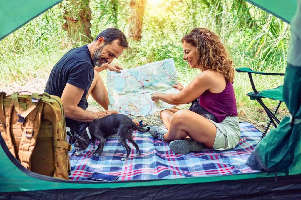 Two people and a dog inside a green tent, looking at a map on a plaid blanket while surrounded by greenery. A backpack is visible beside them, possibly filled with early retirement information as they dream of their plans to retire by 40 amidst nature's beauty.